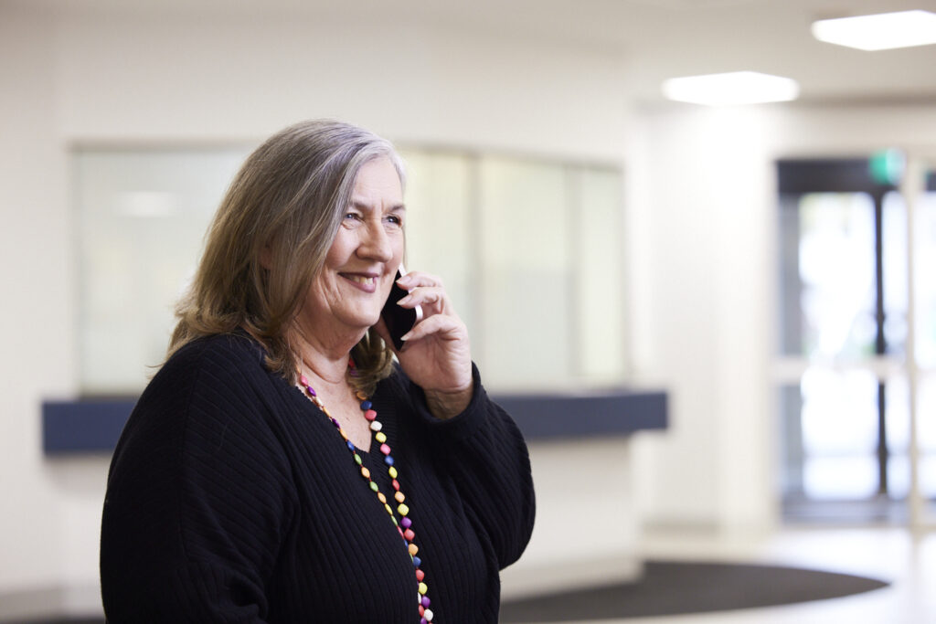 A woman with grey hair stands indoors, smiling while speaking on a mobile phone. She wears a black sweater and a colourful beaded necklace. The background shows a blurred reception area and large windows.
