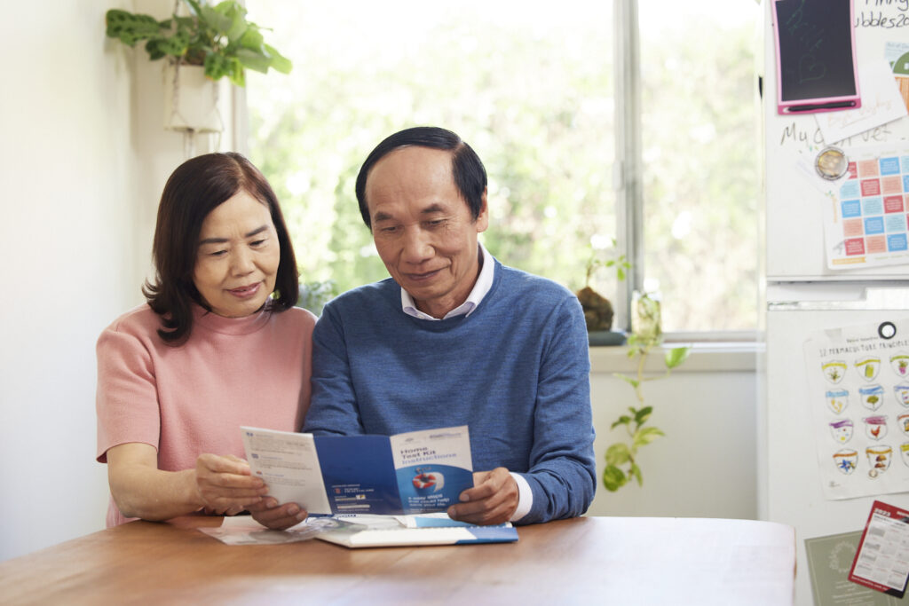 A couple sits together at a kitchen table, reading the instruction booklet for a bowel screening home test kit. The man wears a blue sweater and the woman wears a pink top. A fridge with family magnets and a sunny window are visible in the background.