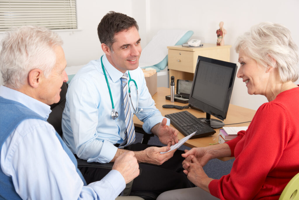 An older couple speaks with a male doctor in a medical office. The doctor, seated behind a desk, holds paperwork and engages them with a friendly expression. Medical equipment and a computer are on the desk.